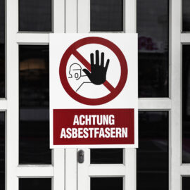 Das Bild zeigt eine weiße Tür mit mehreren rechteckigen Glasfeldern, an der ein großes Warnschild angebracht ist. Das Schild ist überwiegend weiß mit einem roten Kreis und einem schwarzen Symbol, das eine erhobene Hand und eine stilisierte Person mit Schutzmaske darstellt. Unterhalb des Symbols befindet sich ein roter Balken mit dem Text „ACHTUNG ASBESTFASERN“, der auf eine Gefahr durch Asbest hinweist.