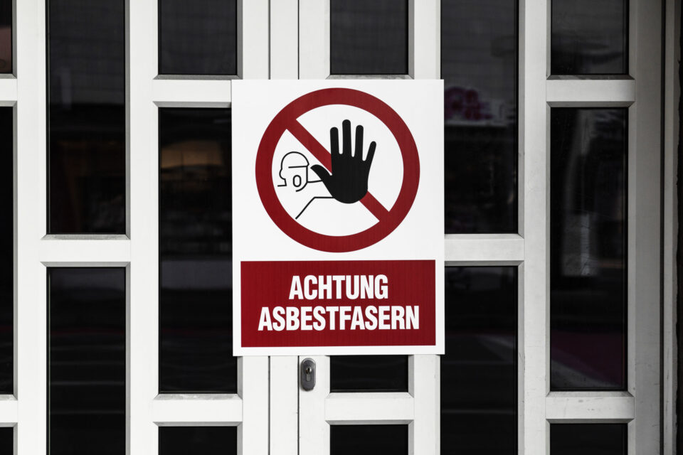 Das Bild zeigt eine weiße Tür mit mehreren rechteckigen Glasfeldern, an der ein großes Warnschild angebracht ist. Das Schild ist überwiegend weiß mit einem roten Kreis und einem schwarzen Symbol, das eine erhobene Hand und eine stilisierte Person mit Schutzmaske darstellt. Unterhalb des Symbols befindet sich ein roter Balken mit dem Text „ACHTUNG ASBESTFASERN“, der auf eine Gefahr durch Asbest hinweist.