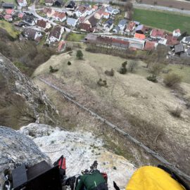 Das Bild zeigt einen steilen Felsen mit einem Schutznetz aus Metall im Vordergrund. Am unteren Bildrand sind Ausrüstungsgegenstände wie Seile und Taschen zu sehen, die auf eine Sicherungs- oder Montagearbeit hindeuten. Im Hintergrund erstreckt sich eine ländliche Landschaft mit Wiesen und einem Dorf mit dicht stehenden Häusern und roten Dächern.