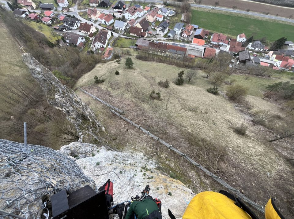 Das Bild zeigt einen steilen Felsen mit einem Schutznetz aus Metall im Vordergrund. Am unteren Bildrand sind Ausrüstungsgegenstände wie Seile und Taschen zu sehen, die auf eine Sicherungs- oder Montagearbeit hindeuten. Im Hintergrund erstreckt sich eine ländliche Landschaft mit Wiesen und einem Dorf mit dicht stehenden Häusern und roten Dächern.