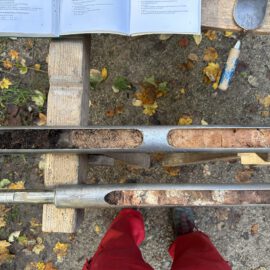 Das Bild zeigt zwei Metallrohre, die auf einer Holzbank liegen und jeweils längliche Aussparungen mit Bodenproben enthalten. Die Proben zeigen unterschiedliche Schichten von dunkler Erde bis hin zu helleren, sandigen und lehmigen Bereichen. Im oberen Bereich liegt ein geöffnetes Buch sowie ein Werkzeug, was auf eine Untersuchung oder Dokumentation der Bodenproben hinweist.