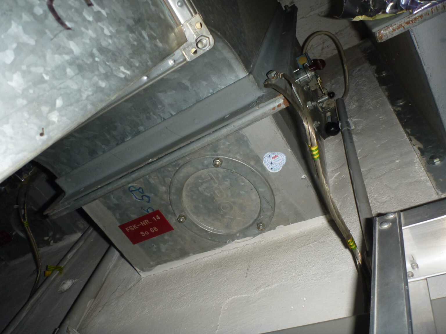 Das Bild zeigt eine metallische Brandschutzklappe, die in ein Lüftungssystem integriert ist. Die Klappe ist rechteckig und mit einer runden Wartungsöffnung versehen, die von Schrauben umgeben ist. An der Seite sind mehrere Kabel und Schläuche angeschlossen, die mit gelben Markierungen versehen sind, und oberhalb verläuft ein verzinkter Lüftungskanal.