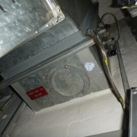 Das Bild zeigt eine metallische Brandschutzklappe, die in ein Lüftungssystem integriert ist. Die Klappe ist rechteckig und mit einer runden Wartungsöffnung versehen, die von Schrauben umgeben ist. An der Seite sind mehrere Kabel und Schläuche angeschlossen, die mit gelben Markierungen versehen sind, und oberhalb verläuft ein verzinkter Lüftungskanal.