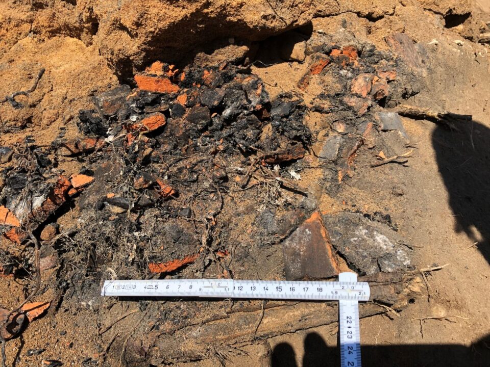 Das Bild zeigt eine Erdschicht mit sandigem Untergrund, in dem zahlreiche dunkle und verbrannte Materialien sowie rote Ziegelbruchstücke liegen. Die dunklen Bestandteile wirken stark verkohlt und sind mit organischen Resten durchsetzt. Im unteren Bereich liegt ein weißer Maßstab mit Zentimeterangaben, der zur Größenbestimmung der Auffüllung dient.