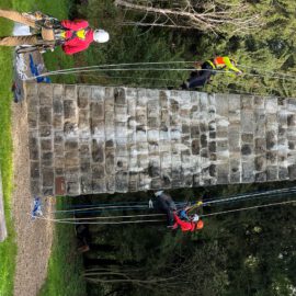 Das Bild zeigt drei Personen, die an einer steinernen Säule im Freien arbeiten und mit Seilen gesichert sind. Zwei Personen hängen an der Säule und steigen mithilfe von Kletterausrüstung und Sicherheitsseilen nach oben, während eine weitere Person am Boden steht und ebenfalls mit Kletterausrüstung ausgestattet ist. Die Umgebung besteht aus einer grünen Wiese, Bäumen im Hintergrund und einem Grillplatz, was auf eine Übung oder Training im Freien hinweist.
