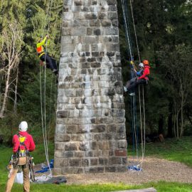 Das Bild zeigt drei Personen, die an einer steinernen Säule im Freien arbeiten und mit Seilen gesichert sind. Zwei Personen hängen an der Säule und steigen mithilfe von Kletterausrüstung und Sicherheitsseilen nach oben, während eine weitere Person am Boden steht und ebenfalls mit Kletterausrüstung ausgestattet ist. Die Umgebung besteht aus einer grünen Wiese, Bäumen im Hintergrund und einem Grillplatz, was auf eine Übung oder Training im Freien hinweist.
