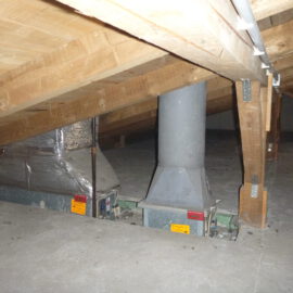 Das Bild zeigt den Dachboden eines Gebäudes mit einer schrägen Holzkonstruktion aus Balken und Metallverbindern. Auf dem Betonboden sind zwei große metallische Lüftungselemente installiert, die mit senkrechten Rohren verbunden sind und teilweise mit silberner Isolierfolie ummantelt sind. Im Hintergrund sind weitere Lüftungskomponenten sichtbar, die ebenfalls in die Dachkonstruktion integriert sind.