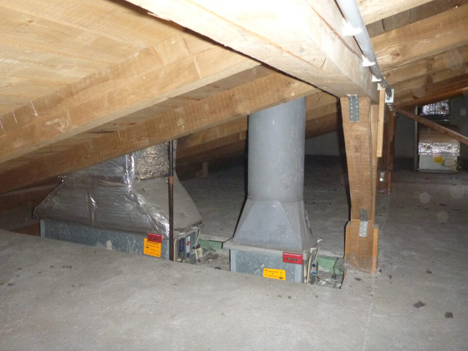 Das Bild zeigt den Dachboden eines Gebäudes mit einer schrägen Holzkonstruktion aus Balken und Metallverbindern. Auf dem Betonboden sind zwei große metallische Lüftungselemente installiert, die mit senkrechten Rohren verbunden sind und teilweise mit silberner Isolierfolie ummantelt sind. Im Hintergrund sind weitere Lüftungskomponenten sichtbar, die ebenfalls in die Dachkonstruktion integriert sind.