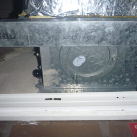 Das Bild zeigt ein metallisches Bauteil, das in eine Zwischendecke eingebaut ist. Es handelt sich um ein rechteckiges Element mit einer runden Öffnung und einem Deckel, der mit Schrauben befestigt ist. Die Umgebung besteht aus einer weißen Rahmenkonstruktion und metallischen Oberflächen, die teilweise mit Folie abgedeckt sind, während die Wand dahinter staubig wirkt.
