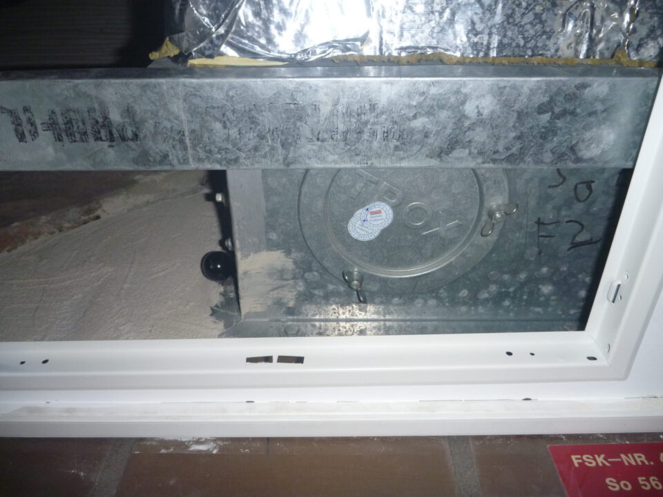 Das Bild zeigt ein metallisches Bauteil, das in eine Zwischendecke eingebaut ist. Es handelt sich um ein rechteckiges Element mit einer runden Öffnung und einem Deckel, der mit Schrauben befestigt ist. Die Umgebung besteht aus einer weißen Rahmenkonstruktion und metallischen Oberflächen, die teilweise mit Folie abgedeckt sind, während die Wand dahinter staubig wirkt.