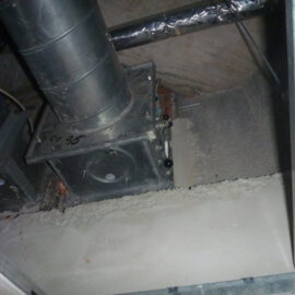 Das Bild zeigt eine geöffnete Zwischendecke mit sichtbarer Lüftungsinstallation. In der Mitte befindet sich ein rechteckiger Metallkasten mit einer runden Revisionsöffnung, aus dem ein senkrechtes Rohr nach oben führt. Weitere Rohre und Leitungen verlaufen im Hintergrund, teilweise mit silberner Isolierung ummantelt.