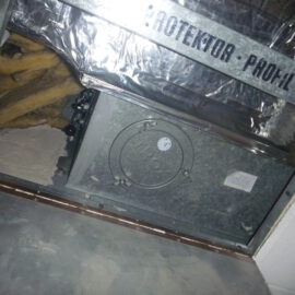 Das Bild zeigt eine geöffnete Zwischendecke mit sichtbarer technischer Installation. In der Mitte befindet sich ein rechteckiger Metallkasten mit einer runden Revisionsöffnung, die mit Schrauben gesichert ist. Darüber sind Dämmmaterialien aus gelber Mineralwolle und eine Folie mit der Aufschrift „Protektor Profil“ zu sehen, die als Abdeckung dient. Die Umgebung wirkt wie ein Bereich für Lüftungs- oder Brandschutztechnik innerhalb eines Gebäudes.