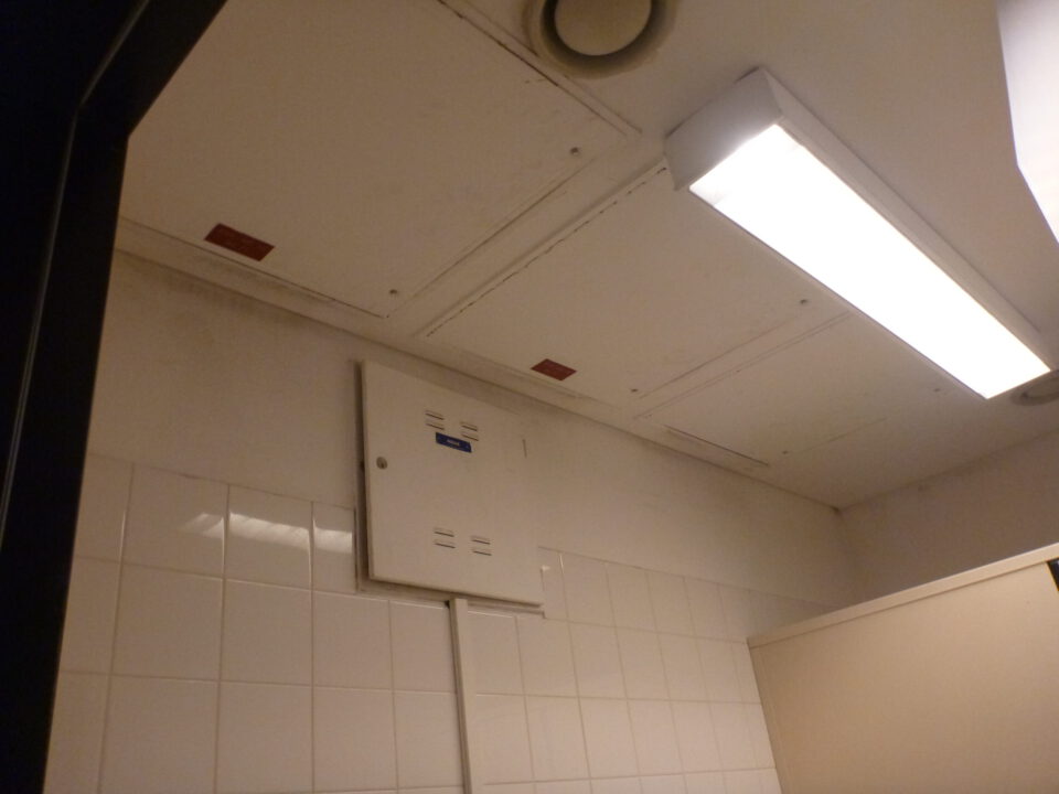Das Bild zeigt die Decke eines Innenraums mit mehreren weißen Revisionsklappen, die in eine Zwischendecke integriert sind. An der Decke ist eine rechteckige Leuchtstofflampe sowie ein runder Lüftungsauslass zu sehen.