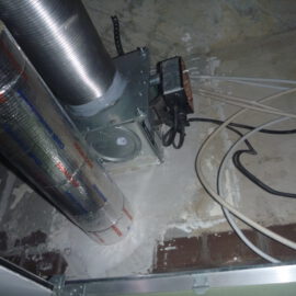 Das Bild zeigt eine geöffnete Zwischendecke mit mehreren technischen Installationen. Im Vordergrund sind zwei senkrechte Rohre zu sehen, von denen eines mit silberner Isolierfolie ummantelt ist und das andere aus blankem Metall besteht. Daneben befindet sich eine metallische Einheit, die mit den Rohren verbunden ist, sowie mehrere lose Kabel in Schwarz und Weiß, die über die staubige Oberfläche verlaufen. Die Umgebung wirkt uneben und zeigt Spuren von Putz und Mauerwerk.