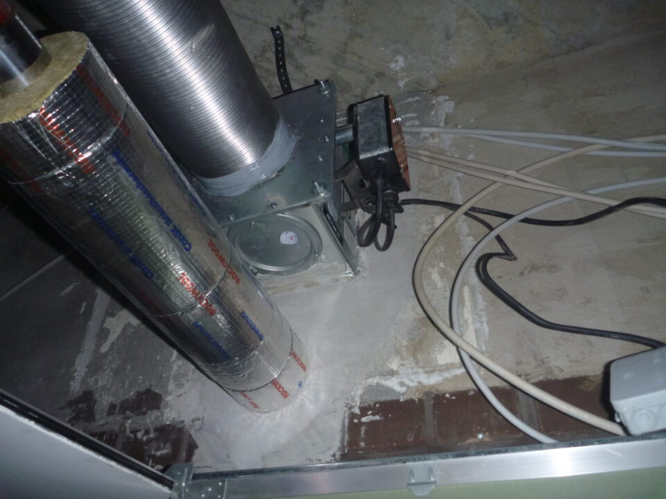 Das Bild zeigt eine geöffnete Zwischendecke mit mehreren technischen Installationen. Im Vordergrund sind zwei senkrechte Rohre zu sehen, von denen eines mit silberner Isolierfolie ummantelt ist und das andere aus blankem Metall besteht. Daneben befindet sich eine metallische Einheit, die mit den Rohren verbunden ist, sowie mehrere lose Kabel in Schwarz und Weiß, die über die staubige Oberfläche verlaufen. Die Umgebung wirkt uneben und zeigt Spuren von Putz und Mauerwerk.