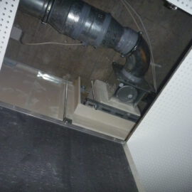 Das Bild zeigt eine geöffnete Zwischendecke mit einem großen schwarzen Rohr, das in einem Bogen verläuft und an einer metallischen Einheit befestigt ist. Die Rohrverbindung ist mit mehreren Metallmanschetten gesichert, und die gesamte Konstruktion wirkt staubig und stark genutzt. Die Umgebung besteht aus einer perforierten weißen Deckenplatte und einem dunklen Bereich im Hintergrund, in dem Kabel und weitere technische Elemente sichtbar sind.