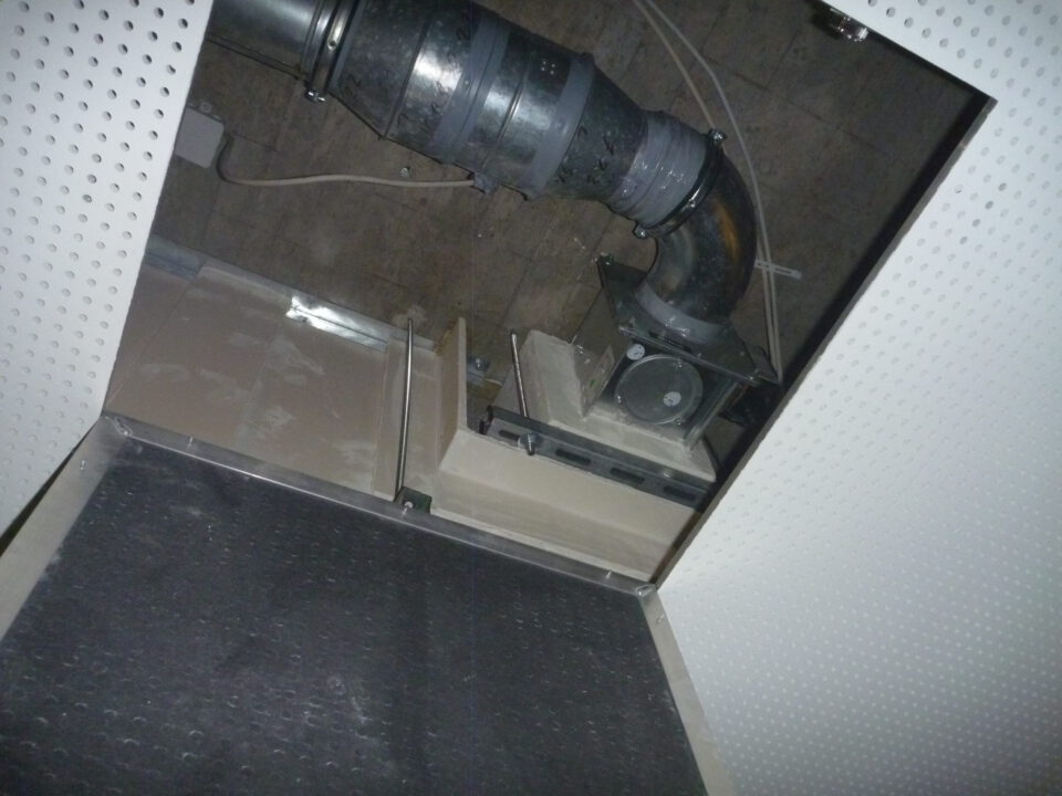 Das Bild zeigt eine geöffnete Zwischendecke mit einem großen schwarzen Rohr, das in einem Bogen verläuft und an einer metallischen Einheit befestigt ist. Die Rohrverbindung ist mit mehreren Metallmanschetten gesichert, und die gesamte Konstruktion wirkt staubig und stark genutzt. Die Umgebung besteht aus einer perforierten weißen Deckenplatte und einem dunklen Bereich im Hintergrund, in dem Kabel und weitere technische Elemente sichtbar sind.