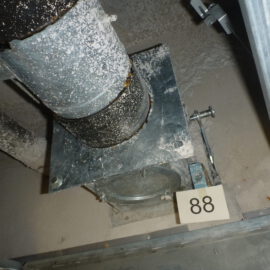 Das Bild zeigt eine geöffnete Zwischendecke mit einem senkrechten Metallrohr, das auf einer rechteckigen Metallplatte befestigt ist. Das Rohr und die Platte sind stark mit Staub und Ablagerungen bedeckt, was auf eine längere Nutzung hinweist. Rechts unten ist eine kleine nummerierte Markierung mit der Zahl 88 zu sehen, und die Umgebung besteht aus grauen, staubigen Flächen sowie weiteren metallischen Bauteilen im Hintergrund.