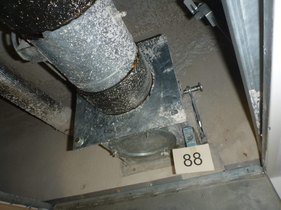 Das Bild zeigt eine geöffnete Zwischendecke mit einem senkrechten Metallrohr, das auf einer rechteckigen Metallplatte befestigt ist. Das Rohr und die Platte sind stark mit Staub und Ablagerungen bedeckt, was auf eine längere Nutzung hinweist. Rechts unten ist eine kleine nummerierte Markierung mit der Zahl 88 zu sehen, und die Umgebung besteht aus grauen, staubigen Flächen sowie weiteren metallischen Bauteilen im Hintergrund.