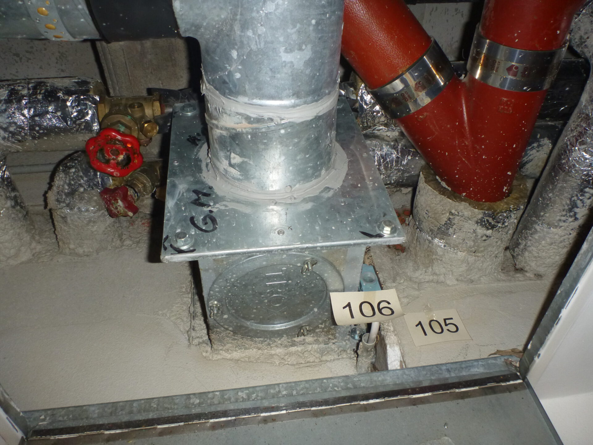 Das Bild zeigt eine geöffnete Zwischendecke mit mehreren sichtbaren Rohrleitungen und technischen Komponenten. In der Mitte befindet sich ein senkrechtes Metallrohr, das auf einer rechteckigen Metallplatte befestigt ist. Rechts verlaufen zwei rote Rohre mit metallischen Rohrschellen, während im Hintergrund weitere Leitungen mit silberner Isolierfolie zu sehen sind. Links ist ein rotes Handrad an einem Ventil erkennbar.