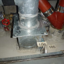 Das Bild zeigt eine geöffnete Zwischendecke mit mehreren sichtbaren Rohrleitungen und technischen Komponenten. In der Mitte befindet sich ein senkrechtes Metallrohr, das auf einer rechteckigen Metallplatte befestigt ist. Rechts verlaufen zwei rote Rohre mit metallischen Rohrschellen, während im Hintergrund weitere Leitungen mit silberner Isolierfolie zu sehen sind. Links ist ein rotes Handrad an einem Ventil erkennbar.