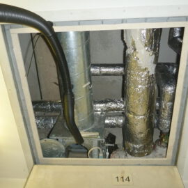 Das Bild zeigt eine geöffnete Zwischendecke mit sichtbaren Installationen. Im Inneren befinden sich mehrere metallische Rohre, von denen einige mit silberner Isolierfolie ummantelt sind, sowie ein senkrechtes Rohr aus blankem Metall. Ein schwarzer flexibler Schlauch verläuft von oben in den Bereich, und am Boden sind Teile einer technischen Einheit zu erkennen.