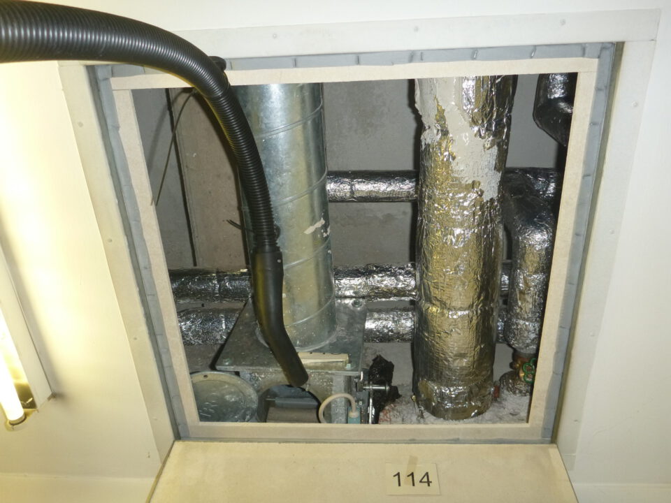 Das Bild zeigt eine geöffnete Zwischendecke mit sichtbaren Installationen. Im Inneren befinden sich mehrere metallische Rohre, von denen einige mit silberner Isolierfolie ummantelt sind, sowie ein senkrechtes Rohr aus blankem Metall. Ein schwarzer flexibler Schlauch verläuft von oben in den Bereich, und am Boden sind Teile einer technischen Einheit zu erkennen.