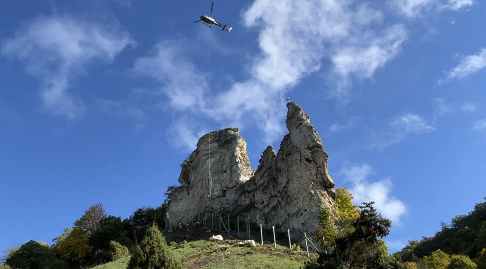 Das Bild zeigt einen Helikopter im Flug über zwei markanten, steilen Felsformationen unter einem blauen Himmel mit vereinzelten weißen Wolken. Vom Helikopter hängt ein langes Seil senkrecht nach unten, das bis zur Spitze der linken Felsnadel reicht. Auf dem Gipfel ist ein kleines Kreuz erkennbar. Unterhalb der Felsen befindet sich ein begrünter Hang mit Sträuchern und Bäumen. Am Fuß der Felsen sind Zäune und einige kleine Figuren von Menschen zu sehen, die sich entlang des Geländes aufhalten.