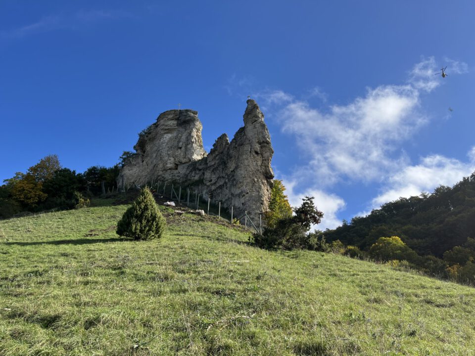 Das Bild zeigt eine markante Felsformation mit zwei hohen, spitzen Felsen, die von einer grünen Wiese und einzelnen Sträuchern umgeben sind. Am unteren Bereich der Felsen ist ein Steinschlagschutzzaun erkennbar, der entlang der Hangkante verläuft. Der Himmel ist klar blau mit einigen weißen Wolken, und rechts oben ist ein Helikopter in der Ferne zu sehen.
