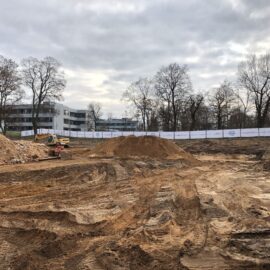 Das Bild zeigt ein freigeräumtes Baufeld mit sandigem Boden und mehreren Erdhügeln. Im Hintergrund sind moderne Gebäude und kahle Bäume zu sehen, die von einem Bauzaun umgeben sind. Auf der Fläche befinden sich deutliche Fahrspuren von Baustellenfahrzeugen, und am linken Bildrand ist ein kleiner Bagger erkennbar.