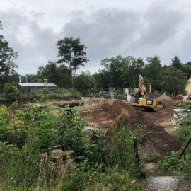 Das Bild zeigt ein Baufeld mit unebenem Gelände, auf dem sich mehrere Erdhügel und offene Flächen befinden. In der Mitte steht ein gelber Bagger auf einem Sandhügel, umgeben von Baumaterialien und Paletten. Im Hintergrund sind Bäume und einige Gebäude zu sehen, während dichter Bewuchs im Vordergrund den Rand der Baustelle markiert.