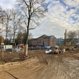 Das Bild zeigt ein weitläufiges Baufeld mit sandigem Boden und deutlichen Fahrspuren von Baustellenfahrzeugen. Mehrere Baumaschinen, darunter Bagger und andere Geräte, sind im mittleren Bereich der Baustelle verteilt, während im Vordergrund ein Fahrzeug teilweise sichtbar ist. Im Hintergrund stehen Gebäude, darunter ein älteres Gebäude mit Gerüst sowie moderne Bauwerke, und die Szene wird von kahlen Bäumen und einem bewölkten Himmel eingerahmt.