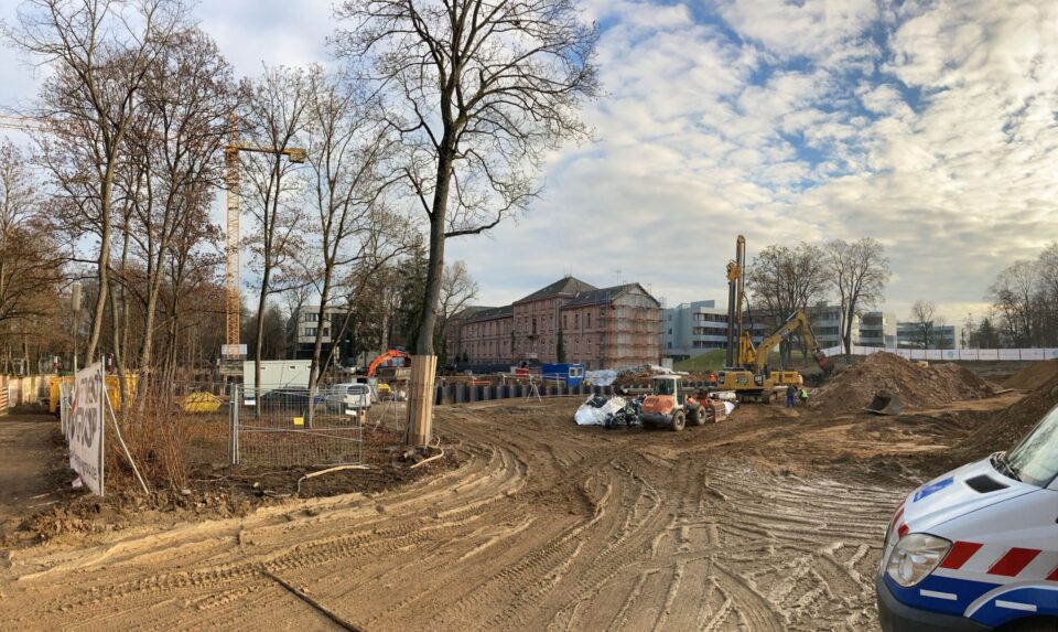 Das Bild zeigt ein weitläufiges Baufeld mit sandigem Boden und deutlichen Fahrspuren von Baustellenfahrzeugen. Mehrere Baumaschinen, darunter Bagger und andere Geräte, sind im mittleren Bereich der Baustelle verteilt, während im Vordergrund ein Fahrzeug teilweise sichtbar ist. Im Hintergrund stehen Gebäude, darunter ein älteres Gebäude mit Gerüst sowie moderne Bauwerke, und die Szene wird von kahlen Bäumen und einem bewölkten Himmel eingerahmt.