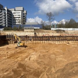 Das Bild zeigt eine große Baugrube mit sandigem Boden, die von einer hohen Spundwand aus Stahl umgeben ist. In der Grube steht ein gelber Bagger, der für Erdarbeiten eingesetzt wird. Im Hintergrund sind ein mehrstöckiges modernes Gebäude, einige Bäume und ein teilweise bewölkter Himmel zu sehen, was auf eine Baustelle in städtischer Umgebung hinweist.