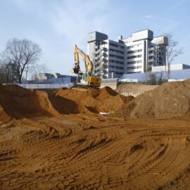 Das Bild zeigt eine große Baugrube mit sandigem Boden, auf dem deutliche Spuren von Fahrzeugketten zu sehen sind. In der Mitte steht ein gelber Bagger auf einem hohen Sandhügel, während im Hintergrund ein mehrstöckiges modernes Gebäude sowie einige Bäume erkennbar sind. Die Szene deutet auf Erdarbeiten im Rahmen einer größeren Baustelle hin.