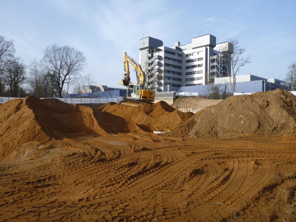 Das Bild zeigt eine große Baugrube mit sandigem Boden, auf dem deutliche Spuren von Fahrzeugketten zu sehen sind. In der Mitte steht ein gelber Bagger auf einem hohen Sandhügel, während im Hintergrund ein mehrstöckiges modernes Gebäude sowie einige Bäume erkennbar sind. Die Szene deutet auf Erdarbeiten im Rahmen einer größeren Baustelle hin.