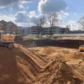 Das Bild zeigt eine große Baugrube mit sandigem Untergrund, in der mehrere hohe Sandhügel aufgeschüttet sind. Zwei große Bagger stehen auf den Sandflächen, einer auf einem Hügel im Vordergrund und der andere weiter hinten in der Grube. Im Hintergrund sind moderne Gebäude, Bäume und ein teilweise bewölkter Himmel zu sehen, was auf eine Baustelle in städtischer Umgebung hinweist.