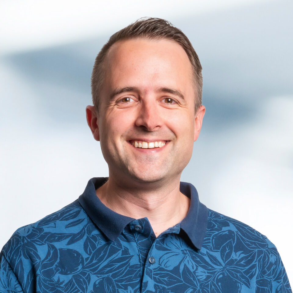 Zu sehen ist ein lächelnder Mann mit kurzem, seitlich gescheiteltem braunem Haar. Er hat ein freundliches, offenes Gesicht und blickt direkt in die Kamera. Er trägt ein blaues Poloshirt mit einem dezenten, floralen Muster.