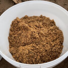 Das Bild zeigt einen weißen Kunststoffeimer, der mit feuchtem, grobkörnigem Sand oder erdähnlichem Material gefüllt ist. Die Oberfläche des Materials ist uneben und leicht verdichtet, was auf eine frisch entnommene Bodenprobe hinweist. Der Eimer steht auf einem erdigen Untergrund, und am Rand sind Teile weiterer weißer Eimer sichtbar.