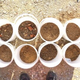 Das Bild zeigt acht weiße Kunststoffeimer, die auf einem kiesigen Untergrund stehen und jeweils mit Bodenmaterial gefüllt sind. Die Inhalte variieren in Farbe und Zusammensetzung, von feinkörnigem Sand bis zu erdigen und tonigen Materialien, teilweise mit kleinen Steinen oder Ziegelbruchstücken. Im unteren Bildbereich sind schwarze Arbeitsschuhe sichtbar, die darauf hindeuten, dass die Aufnahme aus der Perspektive einer stehenden Person gemacht wurde.