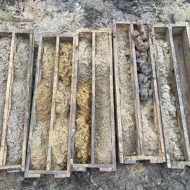Das Bild zeigt mehrere hölzerne Rahmen, die nebeneinander auf dem Boden liegen und jeweils mit Bodenproben gefüllt sind. Die Proben bestehen aus unterschiedlichen Materialien und Farben, von hellen Sand- und Lehmschichten bis hin zu dunkleren, tonigen oder steinigen Bereichen. Die Anordnung deutet auf eine geologische Untersuchung oder eine Analyse von Bohrkernen hin, um die Zusammensetzung des Untergrunds zu dokumentieren.