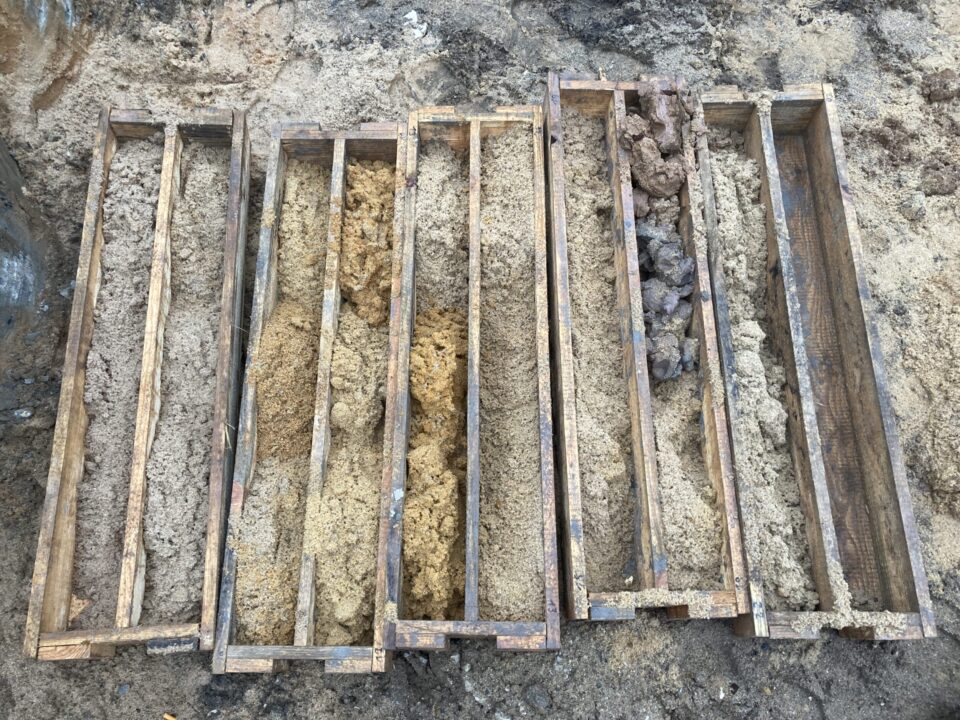 Das Bild zeigt mehrere hölzerne Rahmen, die nebeneinander auf dem Boden liegen und jeweils mit Bodenproben gefüllt sind. Die Proben bestehen aus unterschiedlichen Materialien und Farben, von hellen Sand- und Lehmschichten bis hin zu dunkleren, tonigen oder steinigen Bereichen. Die Anordnung deutet auf eine geologische Untersuchung oder eine Analyse von Bohrkernen hin, um die Zusammensetzung des Untergrunds zu dokumentieren.