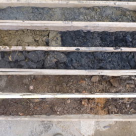 Das Bild zeigt eine Holzkiste mit mehreren Fächern, in denen verschiedene Bodenproben sortiert sind. Die oberen Fächer enthalten graue und dunkelgraue, teilweise klumpige Erde, während die unteren Fächer braune und rötlich-braune Erde mit kleinen Steinen zeigen. Die Anordnung verdeutlicht die unterschiedlichen Schichten, die bei einer Bohrung entnommen wurden.
