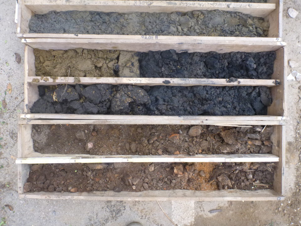 Das Bild zeigt eine Holzkiste mit mehreren Fächern, in denen verschiedene Bodenproben sortiert sind. Die oberen Fächer enthalten graue und dunkelgraue, teilweise klumpige Erde, während die unteren Fächer braune und rötlich-braune Erde mit kleinen Steinen zeigen. Die Anordnung verdeutlicht die unterschiedlichen Schichten, die bei einer Bohrung entnommen wurden.