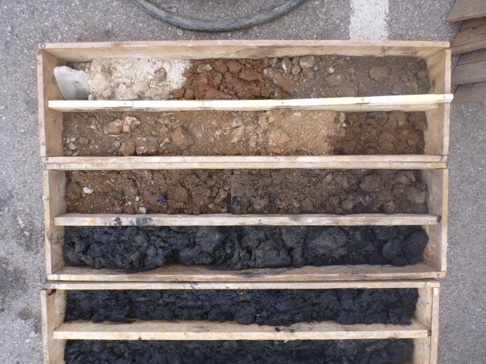 Das Bild zeigt eine Holzkiste mit mehreren Fächern, in denen unterschiedliche Boden- und Gesteinsschichten sortiert sind. Die oberen Fächer enthalten helle und rötliche Steine sowie grobes Material, während die unteren Fächer dunkle, fast schwarze Erde und Gesteinsstücke zeigen. Die Anordnung verdeutlicht die verschiedenen Schichten, die bei einer Bohrung entnommen wurden.