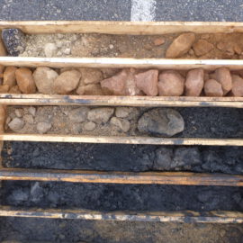 Das Bild zeigt eine Holzkiste mit mehreren Fächern, in denen unterschiedliche Boden- und Gesteinsschichten sortiert sind. Die oberen Fächer enthalten helle und rötliche Steine sowie grobes Material, während die unteren Fächer dunkle, fast schwarze Erde und Gesteinsstücke zeigen. Die Anordnung verdeutlicht die verschiedenen Schichten, die bei einer Bohrung entnommen wurden.