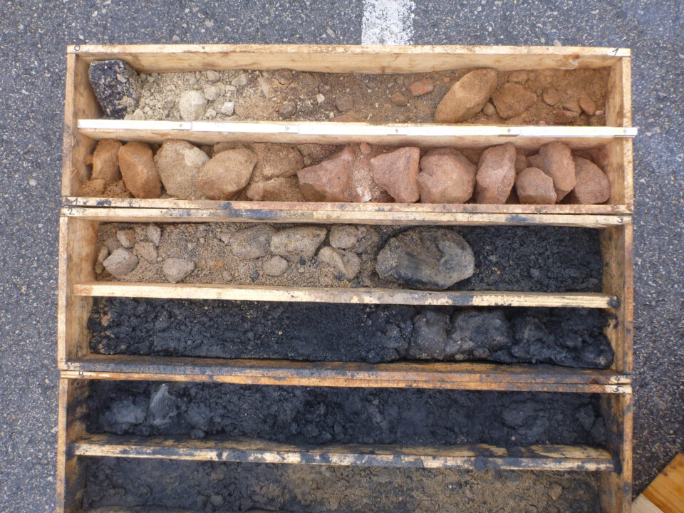 Das Bild zeigt eine Holzkiste mit mehreren Fächern, in denen unterschiedliche Boden- und Gesteinsschichten sortiert sind. Die oberen Fächer enthalten helle und rötliche Steine sowie grobes Material, während die unteren Fächer dunkle, fast schwarze Erde und Gesteinsstücke zeigen. Die Anordnung verdeutlicht die verschiedenen Schichten, die bei einer Bohrung entnommen wurden.