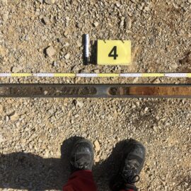Das Bild zeigt eine lange metallische Bohrkernhülse, die auf einem kiesigen Untergrund liegt und mehrere Aussparungen mit Bodenmaterial enthält. Oberhalb der Hülse befindet sich ein gelber Marker mit der Zahl 4 sowie ein Maßstab und ein kleiner zylindrischer Gegenstand zur Größenbestimmung. Im unteren Bereich sind schwarze Arbeitsschuhe sichtbar, was darauf hinweist, dass die Aufnahme aus der Perspektive einer stehenden Person gemacht wurde.