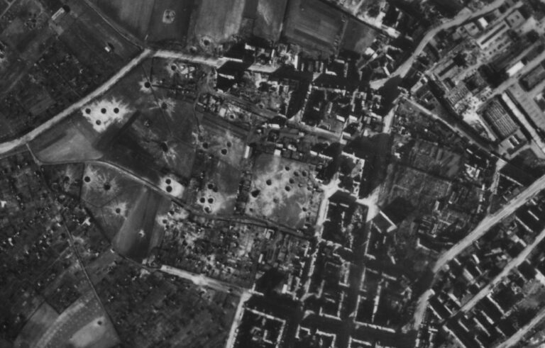 Das Bild zeigt eine historische Luftaufnahme einer Stadt mit deutlich sichtbaren Kriegsschäden. In den offenen Flächen und Feldern sind zahlreiche kreisförmige Krater zu erkennen, die vermutlich durch Bombenexplosionen entstanden sind. Die umliegenden Stadtbereiche bestehen aus dicht angeordneten Gebäuden und Straßen, wobei einige Strukturen beschädigt oder zerstört wirken.