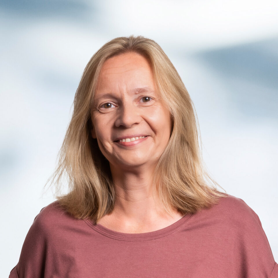 Das Bild zeigt ein professionelles Porträt einer Frau mit schulterlangem, hellblondem Haar, das locker und natürlich fällt. Sie blickt direkt in die Kamera und lächelt freundlich und ruhig. Ihr Gesichtsausdruck wirkt offen, sympathisch und gelassen. Sie trägt ein schlichtes, unifarbenes Oberteil in einem warmen Altrosa-Ton, das einen weichen, harmonischen Kontrast zum Hintergrund bildet.